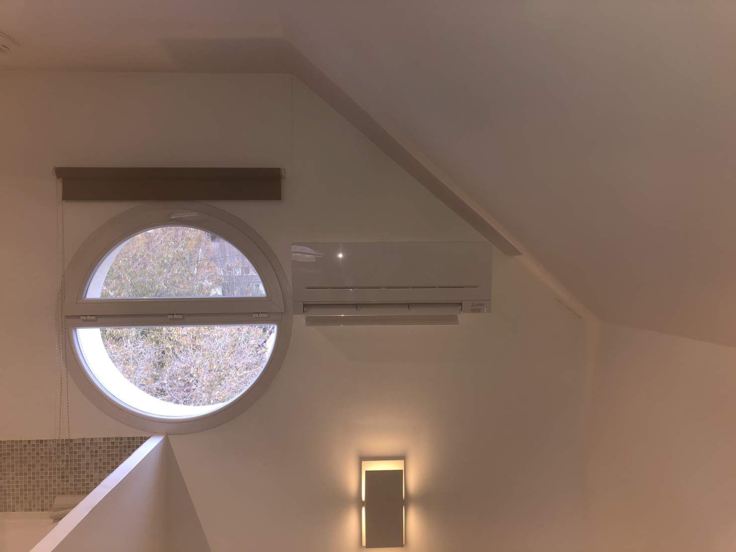 Climatiseur mural réversible posé sous un plafond mansardé, à côté d’une fenêtre ronde
